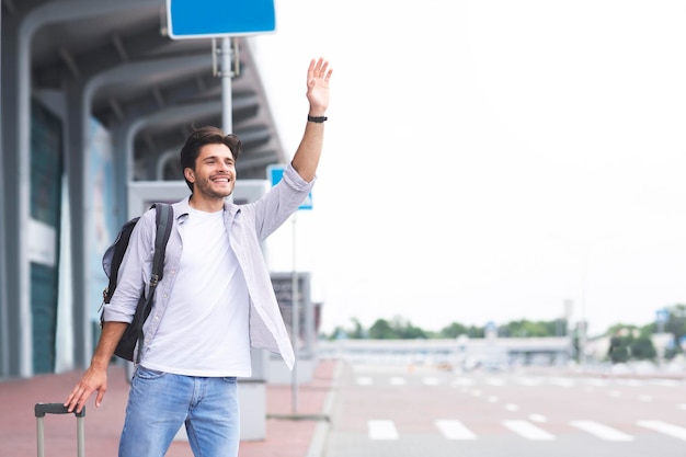 Young guy standing near airport raising hand and trying to stop car