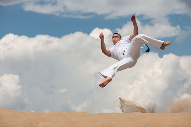 Young guy trains capoeira on sky backround. A men performs martial the kick in the jump