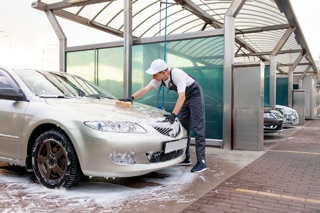 Young guy in uniform car wash worker washes car with sponge with foam man in overalls works