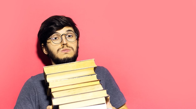 young guy wearing round eyeglasses with bunch of books in hands on coral pink wall.