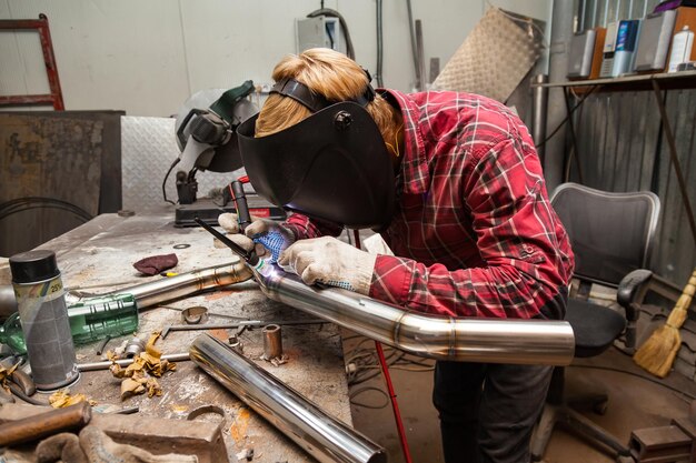 市松模様の赤いシャツを着た若い男性の溶接工が、鉄の作業場でマスクで目を保護するために農業溶接を使用してステンレス鋼のパイプを溶接します。現代の溶接方法