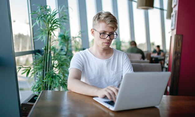 Young guy in a white t-shirt and glasses works on a laptop sitting at a table in a cafeteria