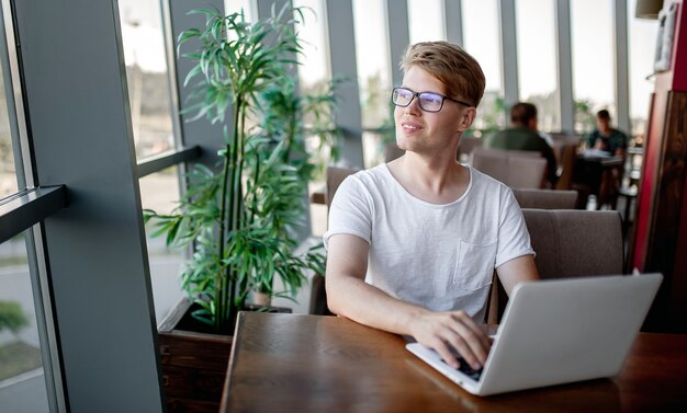 Young guy in a white t-shirt and glasses works on a laptop sitting at a table in a cafeteria