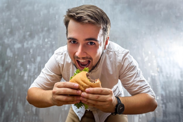 Young guy with a beard eats homemade hamburger on gray background