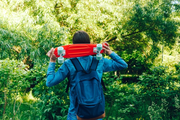 植生の背景を持つ彼の肩に赤いペニーボードを運ぶ青いバックパックを持つ若い男
