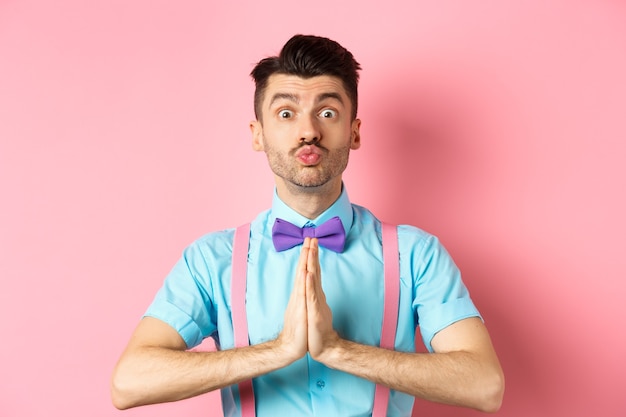 Young guy with cute face pucker lips and asking please, begging for favour, need something and lookin gpleading at camera, standing over pink background.