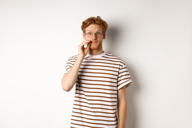 Young guy with ginger hair and glasses zipping mouth, showing lips seal gesture and smiling, keeping a secret, standing over white background.