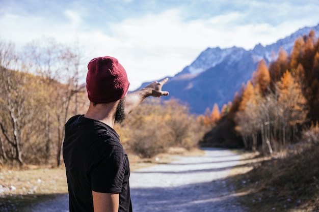 Foto giovane ragazzo con gli occhiali e la barba vestito con una maglietta nera e un cappello rosso che punta alle montagne