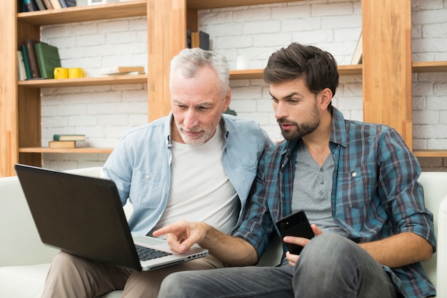 Photo young guy with smartphone pointing at monitor of laptop on legs of aged man on sofa