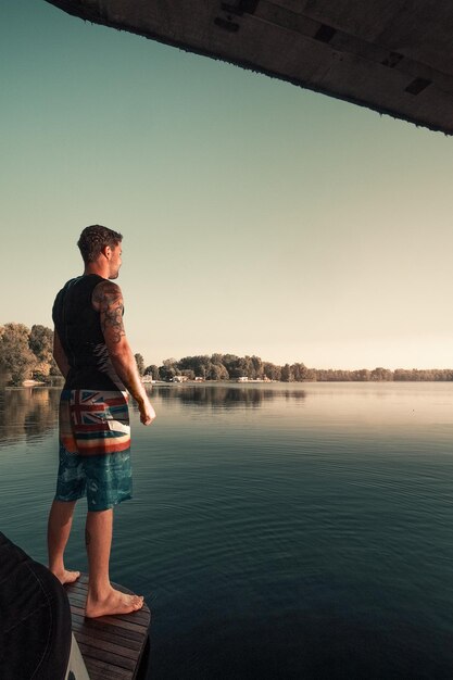 A young guy with tattoos stands on the pier and looks out at the sunrise over the river on summer