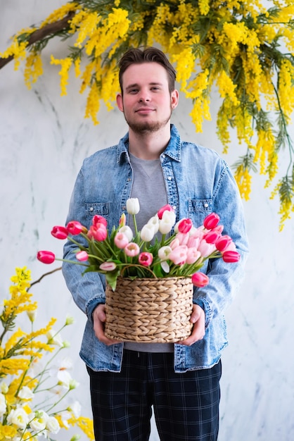 Young guy with a wicker basket of colored tulips in his hands poses against a textured white wall decorated with yellow mimosa