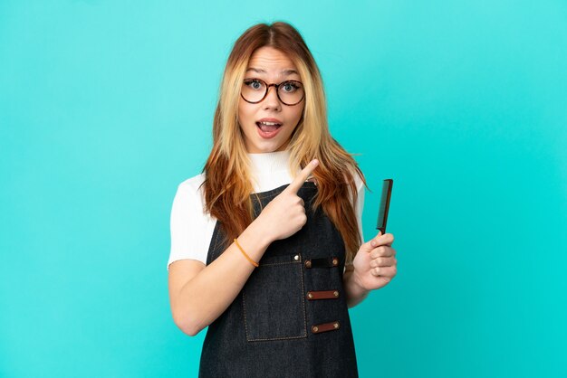 Young hairdresser girl over isolated blue background surprised and pointing side