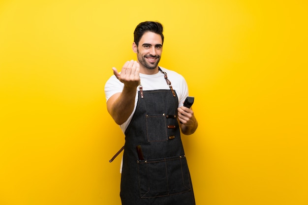 Young hairdresser man over isolated yellow background inviting to come with hand. Happy that you came