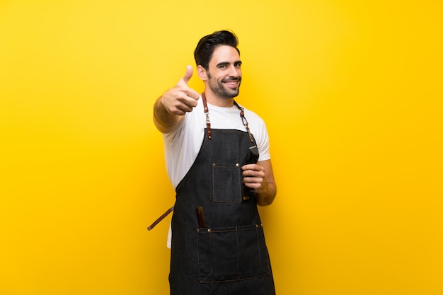 Young hairdresser man over isolated yellow wall with thumbs up because something good has happened