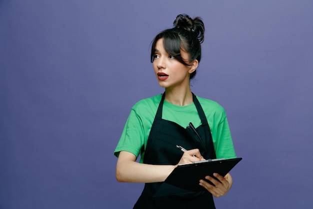 Young hairdresser woman wearing apron holding clipboard and pen looking aside with serious face making notes standing over blue background