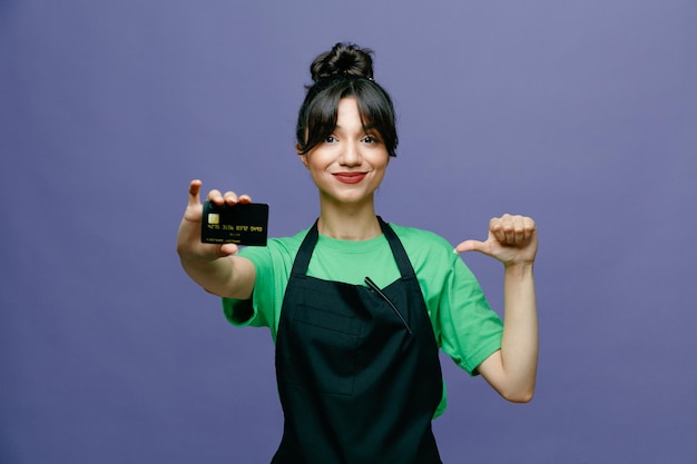 Young hairdresser woman wearing apron holding credit card looking at camera smiling confident pointing with thumb at herself standing over blue background