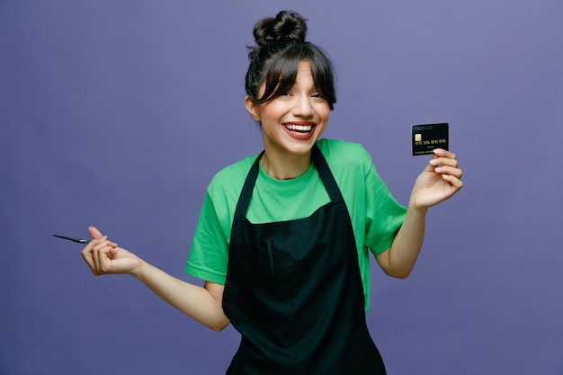 Young hairdresser woman wearing apron holding credit card and scissors looking at camera happy and positive smiling cheerfully standing over blue background