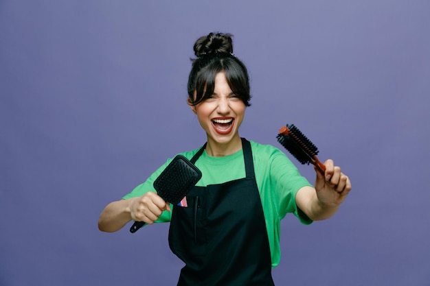 Young hairdresser woman wearing apron holding hair brushes looking at camera happy and joyful smiling cheerfully standing over blue background