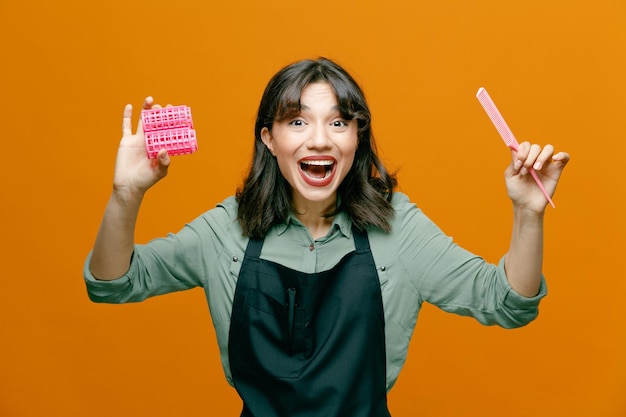 Photo young hairdresser woman wearing apron holding hair comb hair curlers looking at camera happy and positive smiling cheerfully standing over orange background