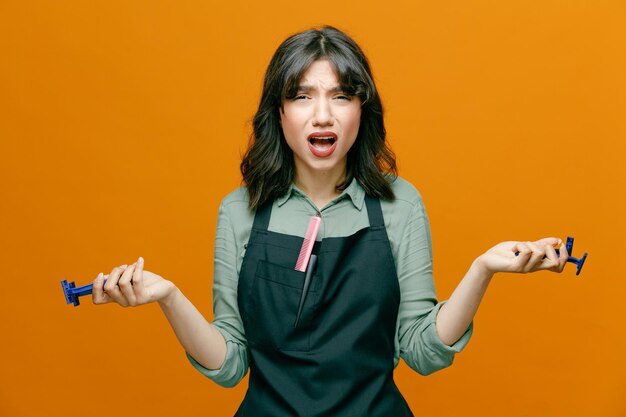 young hairdresser woman wearing apron holding razors looking at camera confused and disappointed standing over orange background