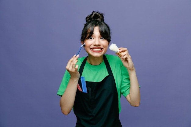 Young hairdresser woman wearing apron holding shaving brush and razor looking at camera happy and joyful smiling broadly standing over blue background