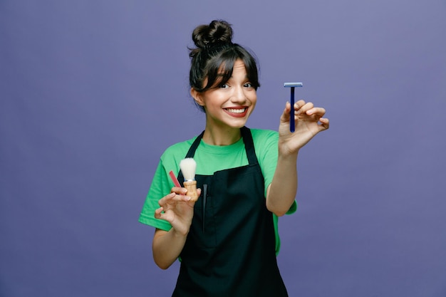 Young hairdresser woman wearing apron holding shaving brush and razor looking at camera happy and positive smiling cheerfully standing over blue background