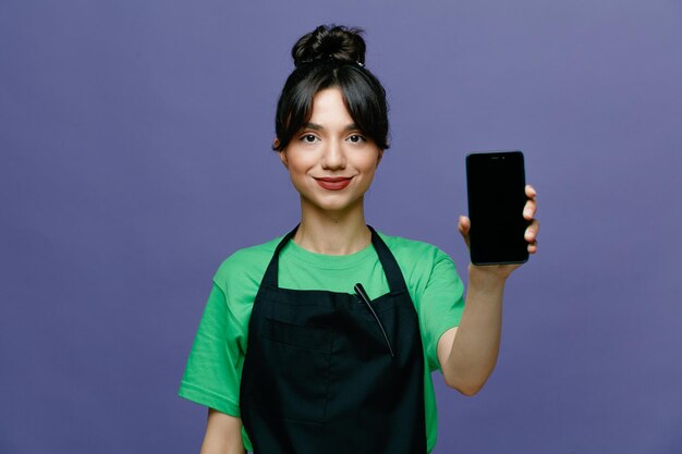 Young hairdresser woman wearing apron showing smartphone looking at camera smiling confident standing over blue background