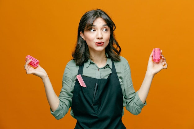 Young hairdresser woman wearing apron with comb holding hair curlers looking aside surprised with confuse expression standing over orange background