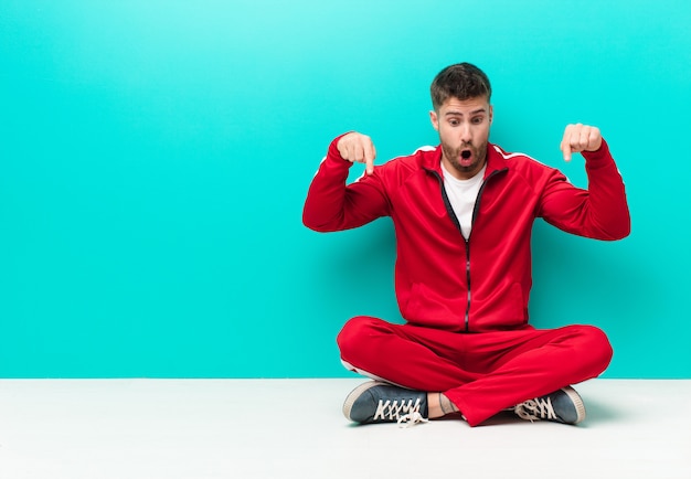 Young handosme man feeling shocked, open-mouthed and amazed, looking and pointing downwards in disbelief and surprise against flat color wall