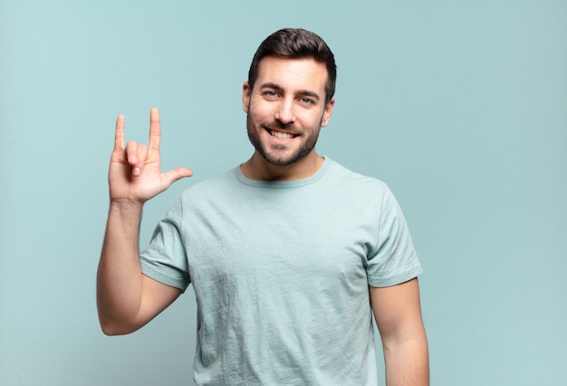 Young handsome adult man feeling happy, fun, confident, positive and rebellious, making rock or heavy metal sign with hand