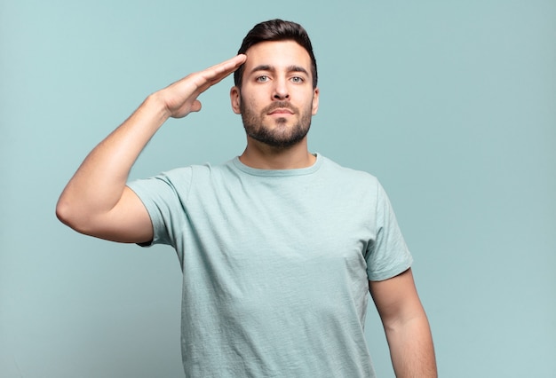 Young handsome adult man greeting the camera with a military salute in an act of honor and patriotism, showing respect