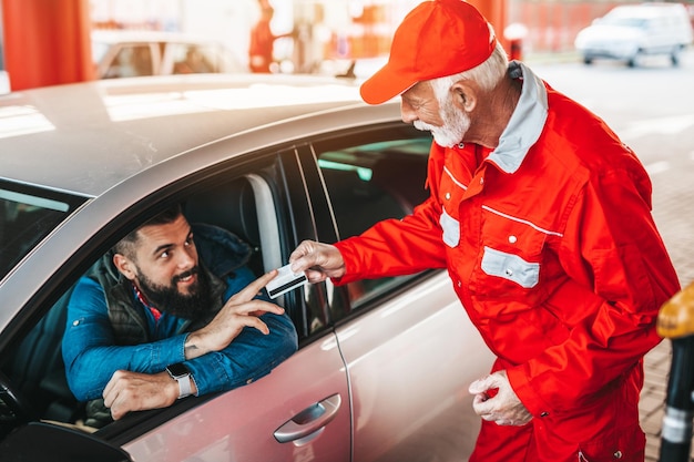 Young handsome adult man together with senior worker standing on gas station and fueling car and paying with credit card.