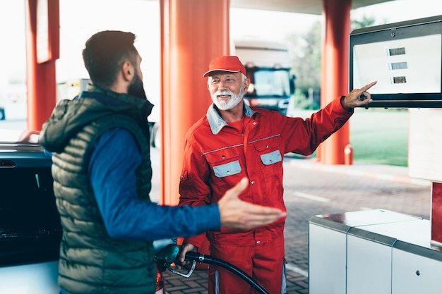 ガソリンスタンドに立って車に燃料を補給するシニアワーカーと一緒に若いハンサムな大人の男。