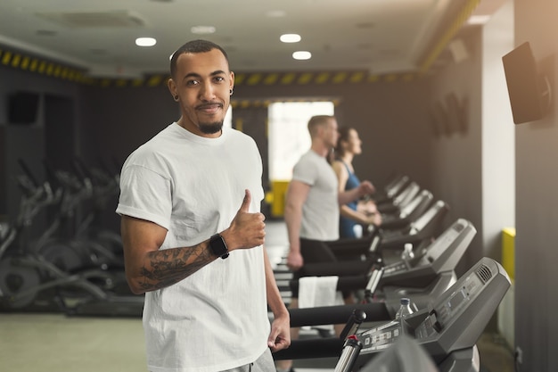 Young handsome african-american man in fitness club, showing thumb up and smiling at camera. Cardio workout, running on treadmill. Healthy lifestyle, guy training in gym, copy space