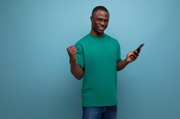 Молодой красивый африканский парень с короткой стрижкой, одетый в простую футболку, использует смартфон для