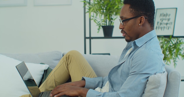 Young handsome african man in glasses use laptop sitting on sofa work at home online technology internet