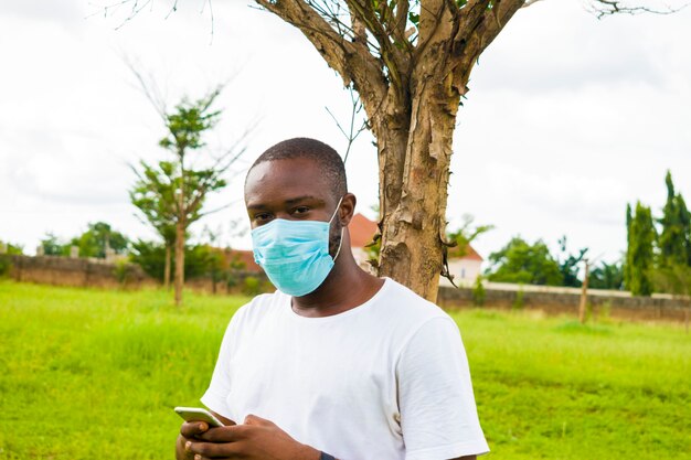 Giovane bell'uomo africano seduto fuori indossando una maschera facciale per prevenire, prevenire, prevenire lo scoppio nella società mentre usa il suo cellulare