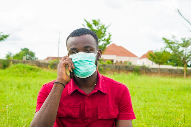 Giovane uomo africano bello che indossa la maschera per il viso che fa chiamate con il suo cellulare.