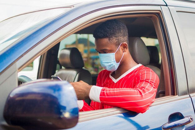 Il giovane tassista africano bello che indossa la maschera facciale che previene, previene, si impedisce lo scoppio nella società e gestisce il suo telefono cellulare