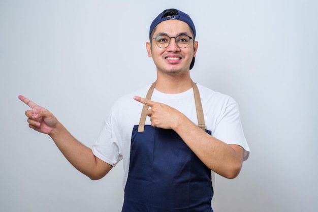 Young handsome asian barista man wearing apron pointing something on his side with his both hand