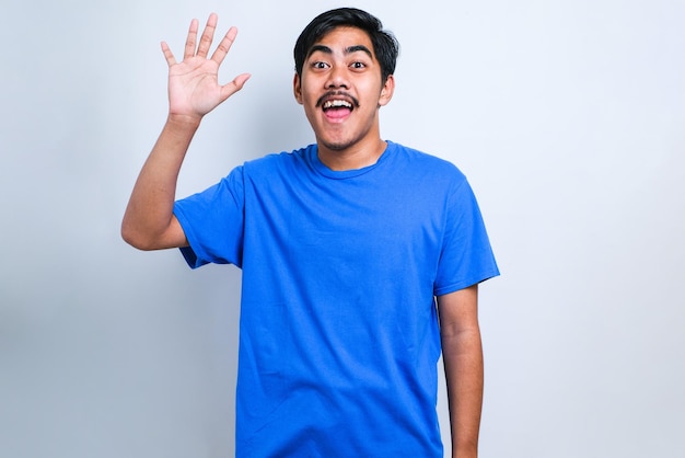 Young handsome asian boy wearing white tshirt standing over white background Waiving saying hello happy and smiling, friendly welcome gesture