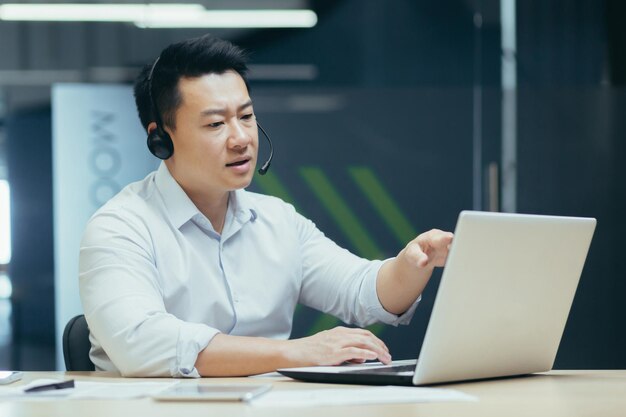 A young handsome asian businessman in headphones with a microphone sits at a laptop in the office