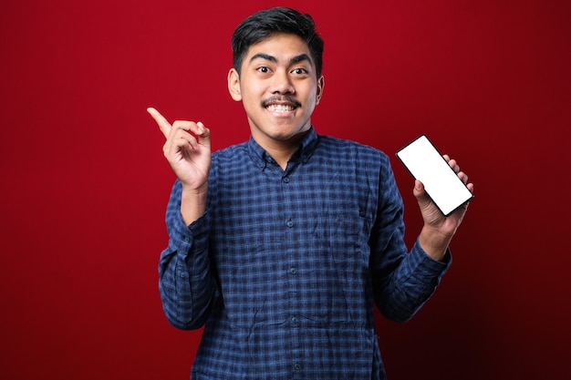 Young handsome asian man showing screen of smartphone over red isolated background very happy pointing with hand and finger to the side