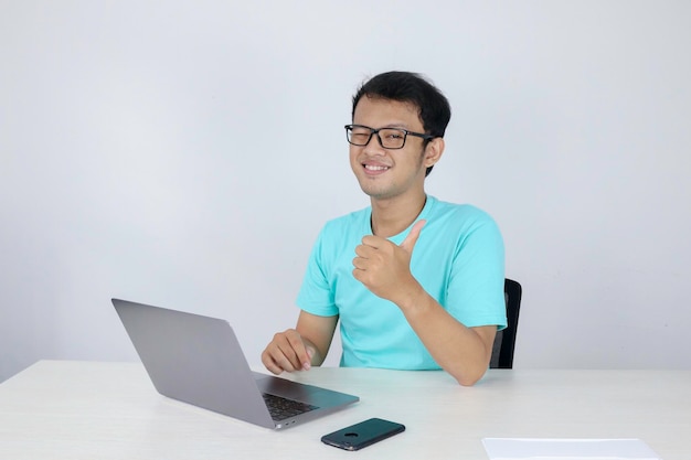 Young handsome asian man showing thumb up while working with laptop Indonesian man wearing blue shirt