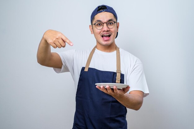 Young handsome asian man wearing apron showing excited expression while holding empty dinner plate