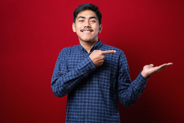 Young handsome asian man wearing casual flannel shirt over red background amazed and smiling to the camera while presenting with hand and pointing with finger.