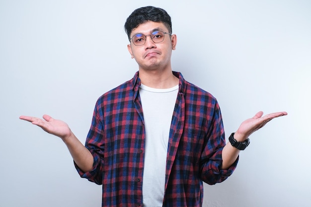 Young handsome Asian man wearing casual shirt and glasses cueless and confused expression with arms and hands raised