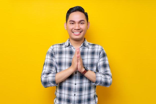 격자무늬 셔츠를 입은 젊고 잘생긴 아시아 남자는 노란색 배경에 고립된 큰 미소로 인사하는 손을 줍니다.