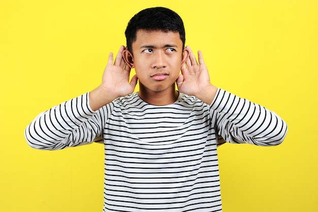 Молодой красивый азиатский мужчина в полосатой футболке слушает что-то рукой над ухом, слушая слухи или сплетни. Понятие глухоты