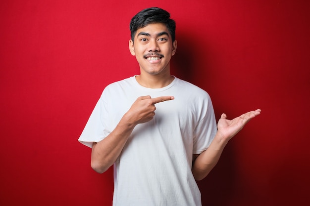 Young handsome asian man wearing white t-shirt over red background amazed and smiling to the camera while presenting with hand and pointing with finger.